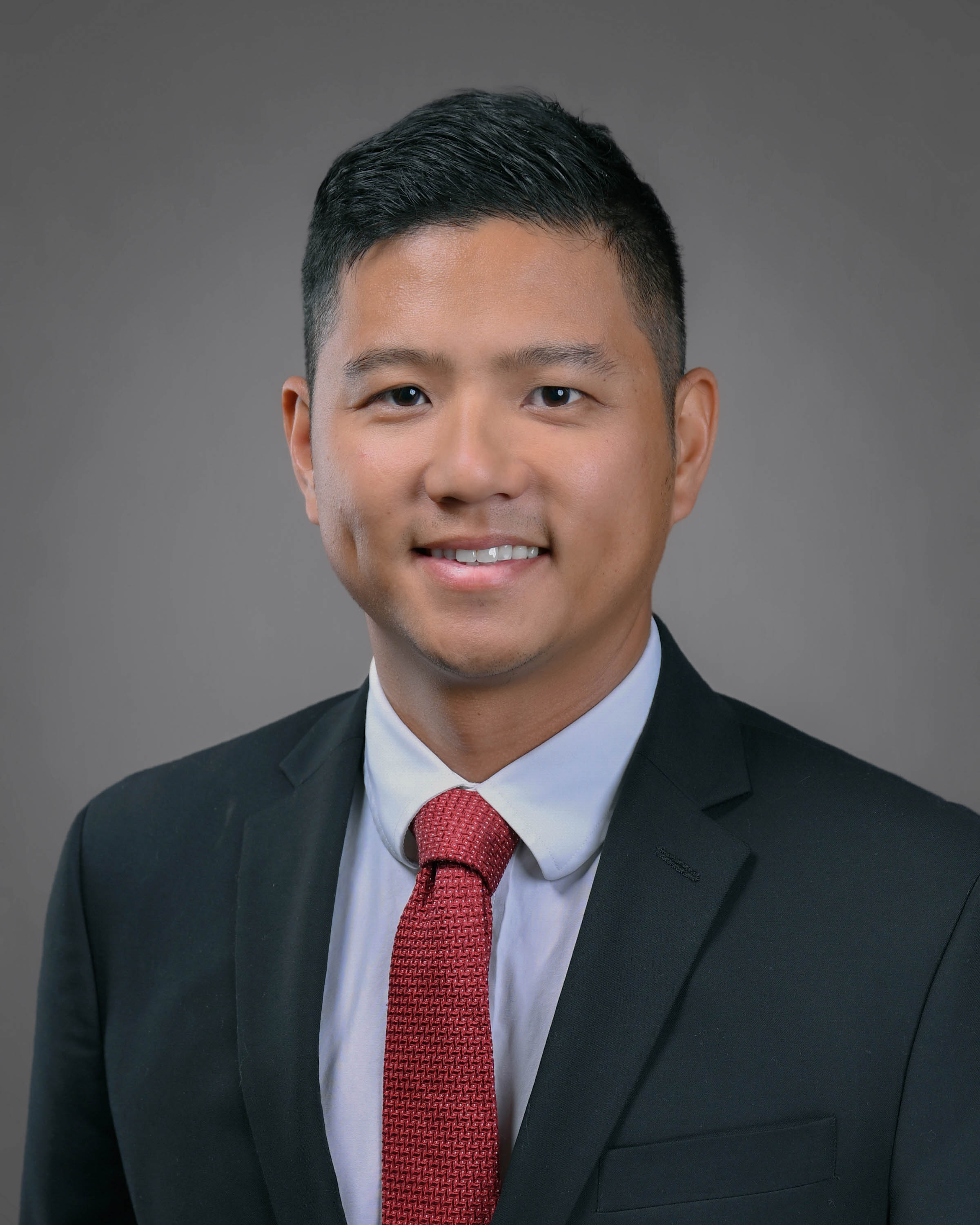 Man with short dark hair smilng in a dark suit with red tie